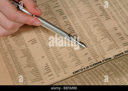 Mano che tiene una penna argento per controllare i prezzi del mercato mondiale da un quotidiano finanziario pagina Foto Stock