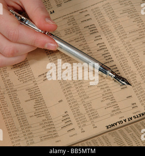 Mano che tiene una penna argento per controllare i prezzi del mercato mondiale da un quotidiano finanziario pagina Foto Stock