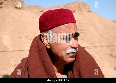 Il vecchio uomo con un baffi grigio indossando un tradizionale djellaba e red fez in Chenini, Tunisia Foto Stock