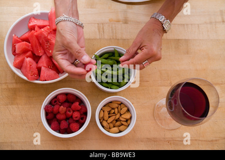 Primo piano delle mani con ciotole di articoli alimentari su un blocco di trinciatura compresi anguria lamponi fagioli di soia edamame mandorle. Foto Stock