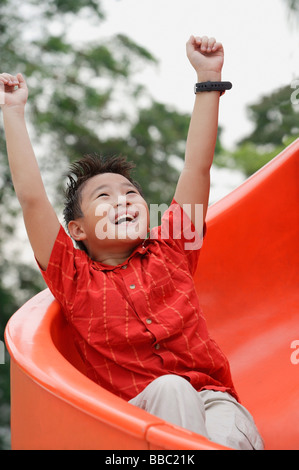 Ragazzo scendendo parco giochi diapositiva, braccia tese, sorridente, guardando verso l'alto Foto Stock