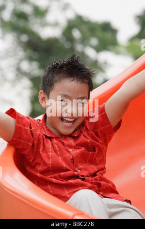 Ragazzo scendendo parco giochi diapositiva, braccia tese, sorridente Foto Stock