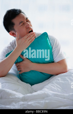 Uomo seduto nel letto, abbracciando cuscino, sbadigli Foto Stock