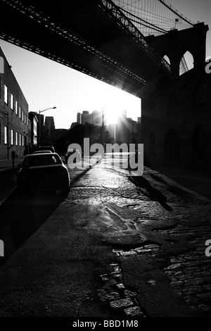 Sun riflettendo in acciottolato di una Street in Brooklyn con un motociclo silhouette sotto il ponte di Brooklyn a New York Foto Stock