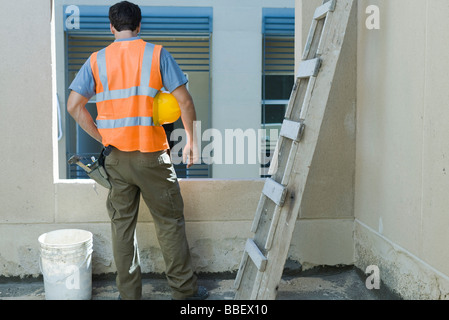 Lavoratore edile con mano sul hip a guardare fuori dalla finestra, vista posteriore Foto Stock