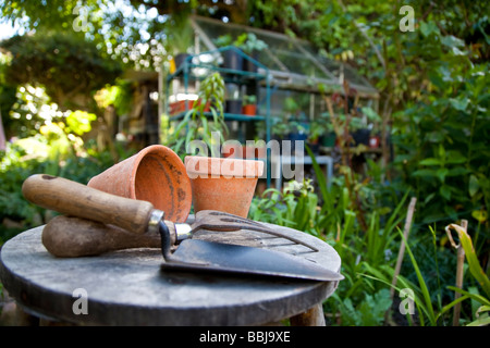 Utensili da giardinaggio e vasi di fiori appoggiati su uno sgabello in un giardino verde con una serra al di fuori della messa a fuoco in background Foto Stock