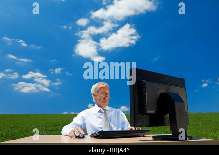 Il concetto di Business shot che mostra un vecchio Esecutivo maschio utilizzando un computer in un campo verde con un cielo blu e soffici nuvole bianche Foto Stock