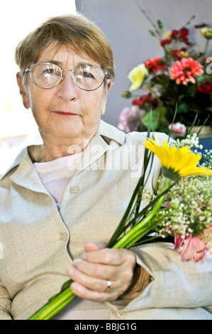Donna anziana con gli occhiali azienda fiori e sorridente Foto Stock