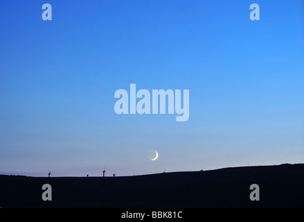 Silver crescent moon nel naufragio di un blu, twilight sky west di un moortop della silhouette Ovenden Moor Wind Farm, Halifax, Regno Unito Foto Stock