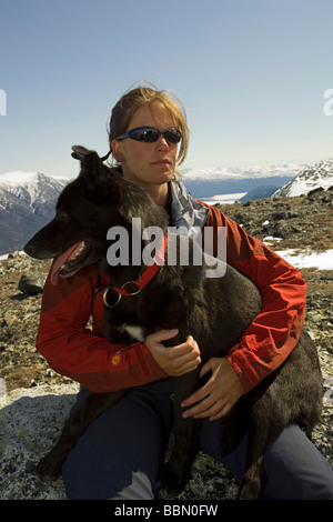 Escursionista in appoggio, seduto su una roccia, giovane donna con il cane, sled dog, Alaskan husky, montagne dietro, Kusawa Ridge, Yukon Territor Foto Stock