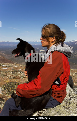 Escursionista in appoggio, seduto su una roccia, giovane donna con il cane, sled dog, Alaskan husky, montagne dietro, Kusawa Ridge, Yukon Territor Foto Stock