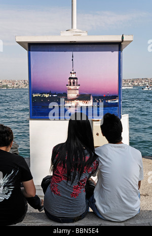 Coppia giovane seduto sulla riva del Bosforo, guardando un poster atmosferica della torre di Leandro su un capannone, Kabatas, Istanbul Foto Stock