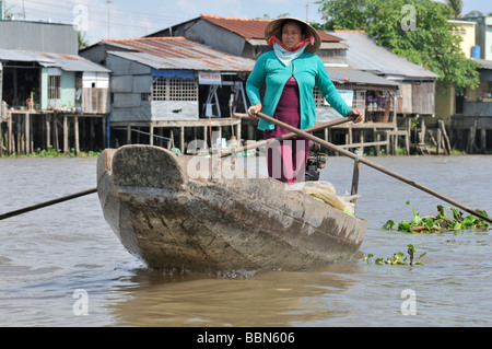 Donna con cappello tradizionale a forma di cono hat fatte di foglie di palma, permanente e la barca a remi di una barca di legno sul Mekong, carico di frutta Foto Stock