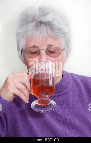 Anziana donna senior pensionati lady indossando un ponticello di porpora e di bere frutta rossa tè in una tazza di vetro. Inghilterra Regno Unito Gran Bretagna Foto Stock