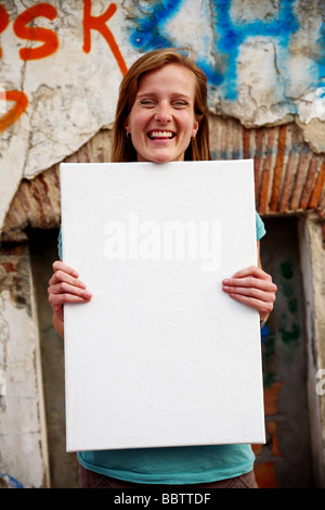 Giovane donna bianca tenendo vuoto bianco segno di tela nella parte anteriore del vecchio muro con graffiti. Foto Stock
