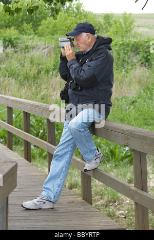 I vecchi maturo maschio bianco seduto sulla ringhiera in legno in giacca e jeans e ballcap riprese video digitali in ambiente esterno Foto Stock