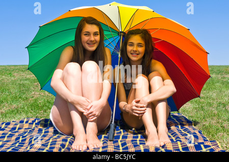 Ritratto orizzontale di due giovani sorelle stessi di schermatura dal sole sotto un enorme ombrello coloratissimo su un giorno d'estate Foto Stock
