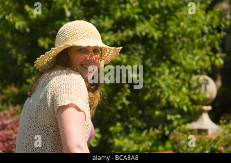 Giovane donna in un cappello sorridente e guardandosi le spalle contro un fuori fuoco sullo sfondo del giardino Foto Stock