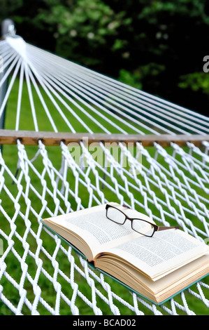 Lettura estiva staycation home vacanze weekend di pensionamento concetto occhiali occhiali occhiali libro aperto backyard amaca. Foto Stock