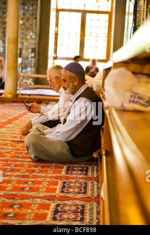 La Turchia , Istanbul , interno nuova moschea o Yeni Cami , uomo barbuto in cap pregando su tappeti mentre uno sul cellulare o telefono cellulare Foto Stock
