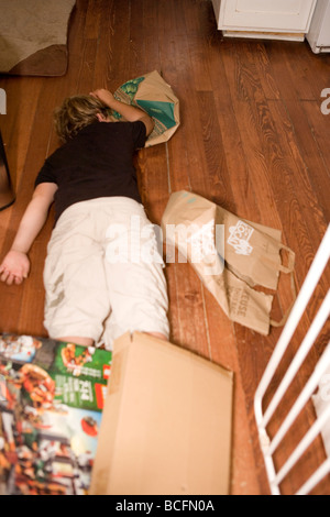 Dieci anni di vecchio ragazzo che stabilisce sul pavimento a casa stanchi dopo aver giocato tutto il giorno, circondato da scatole di cartone Foto Stock