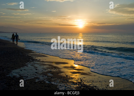 L uomo e la donna a piedi lungo una spiaggia tenendo le mani al tramonto Adelianos Kampos Foto Stock