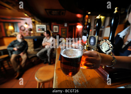 Due uomini seduti a un fuoco aperto in un pub tradizionale come la padrona di casa mette una pinta di birra al bar GLOUCESTERSHIRE England Regno Unito Foto Stock