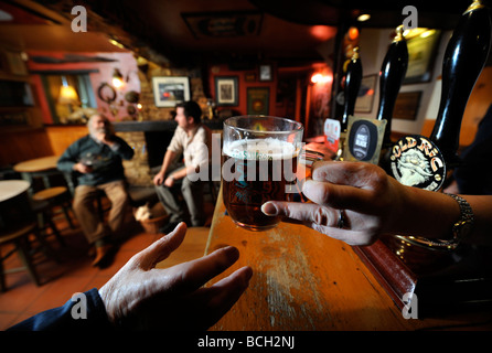 Due uomini seduti a un fuoco aperto in un pub tradizionale come la padrona di casa serve una pinta di birra al di sopra del bar GLOUCESTERSHIRE England Regno Unito Foto Stock