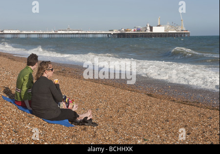 Due vacanzieri seduti sulla spiaggia di ciottoli vicino al mare e le onde del mare di fronte al molo di Brighton come marea avanza. Foto Stock