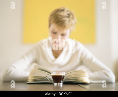 Donna seduta al tavolo al libro di lettura, una tazza di caffè in primo piano Foto Stock