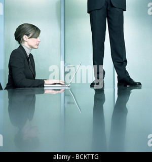 Donna seduta al tavolo al lavoro su laptop, uomo in piedi sul tavolo Foto Stock