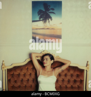 Donna seduta su un divano con le mani dietro la testa e gli occhi chiusi, sotto foto tropicale sulla parete Foto Stock