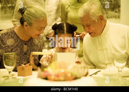 Nonni e nipote apertura regalo di compleanno, seduti fianco a fianco a tavola Foto Stock