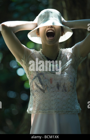 Giovane donna in piedi all'aperto, indossando il cappello sopra gli occhi, gridando, a braccia alzate Foto Stock