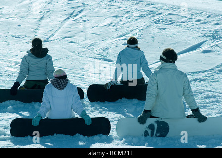 Quattro giovani snowboarders seduti sulla pista da sci, vista posteriore Foto Stock