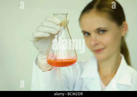 Donna scienziato azienda fino in bicchiere con liquido interno, guardando la fotocamera, la messa a fuoco su oggetti in primo piano Foto Stock
