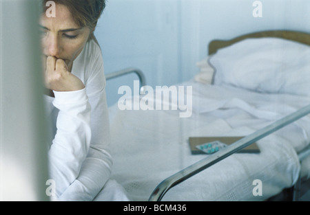 Donna seduta sul letto di ospedale, guardando verso il basso con la mano sotto il suo mento, vista ritagliata Foto Stock