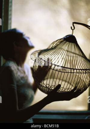 Donna in piedi accanto alla finestra, tenendo vuoto birdcage Foto Stock