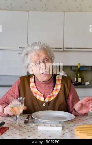 Frau in ihren Siebzigern sitzt in der Küche am gedeckten Mittagstisch Foto Stock