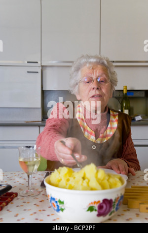 Frau in ihren Siebzigern sitzt in der Küche am gedeckten Mittagstisch und nimmt sich Kartoffeln auf Ihren Teller Foto Stock