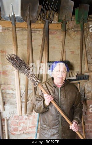 Frau in ihren Siebzigern mit Kopftuch steht vor einer Wand mit Werkzeugen und hält einen Reisigbesen Foto Stock