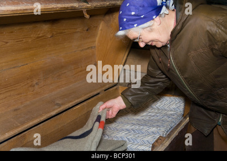 Frau in ihren Siebzigern mit Kopftuch schaut in Eine große alte Wäschetruhe Foto Stock
