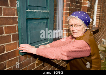 Frau in ihren Siebzigern mit Kopftuch schließt die Fensterläden eines Hauses Foto Stock