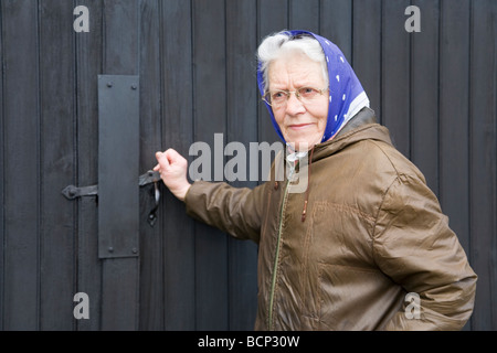 Frau in ihren Siebzigern mit Kopftuch schließt den Riegel am Scheunentor auf ihrem Bauernhof Foto Stock