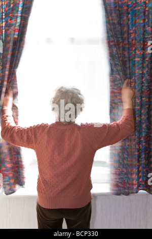 Frau in ihren Siebzigern steht morgens am Fenster und zieht die Gardinen zur Seite Foto Stock