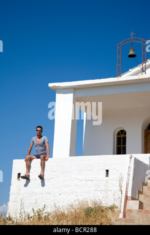 Uomo in pantaloncini e maglietta siede sulla cappella bianca parete in Grecia Foto Stock