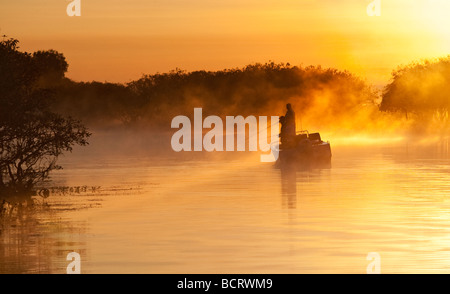 Il sole che sorge dietro una barca da pesca mentre un inizio di mattina nebbia derive nel acqua gialla billabong nel Parco Nazionale Kakadu, Aus Foto Stock