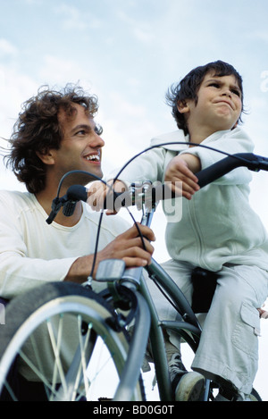 Ragazzino sulla bicicletta si ostina a guardare lontano, padre inginocchiato a lato rassicurante sorridente Foto Stock