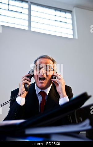 Manager con gli occhiali e tuta seduto di fronte a una montagna di cartelle, rabbiosamente parlando a due linee telefoniche Foto Stock