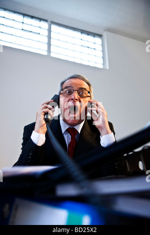 Manager con gli occhiali e tuta seduto di fronte a una montagna di cartelle, rabbiosamente parlando a due linee telefoniche Foto Stock